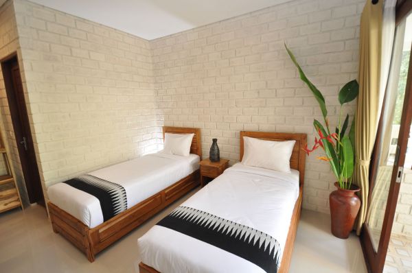 Cozy Bali-style twin bedroom with wooden beds, crisp white linens, tropical heliconia plant, and exposed light brick walls.